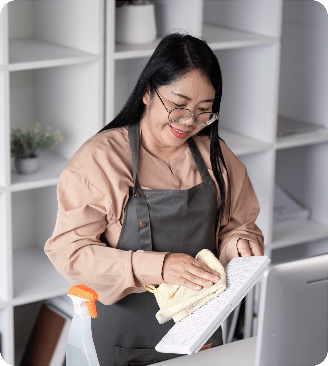 Woman in apron cleans keyboard with cloth in bright office; spray bottle nearby